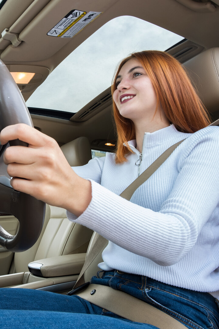 Wide angle view of young redhead woman driver fastened by seatbelt driving a car smiling happily